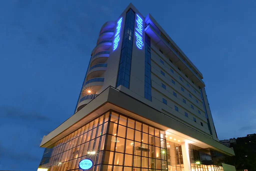 Fachada do Agil Hotel ao anoitecer, com o letreiro azul aceso no alto do prédio